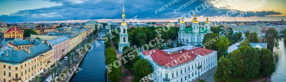 Панорамная фотография летнего Санкт-Петербурга с высоты птичьего полета. На снимке виден Никольский Морской собор, Крюков канал и окружающие исторические здания центра города. Фотография передает атмосферу солнечного дня, отражения воды в канале и гармоничное сочетание архитектуры и городской среды Петербурга. Вид с дрона позволяет оценить масштабы застройки, красоту архитектурных ансамблей и уникальные панорамные виды Северной столицы России. Этот снимок подходит для туристических публикаций, фотостоков, оформления интерьеров, рекламы и проектов, связанных с городской архитектурой и культурным наследием Петербурга.